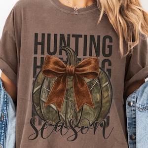 May include: Brown t-shirt with a pumpkin design. The pumpkin has a camouflage pattern and a large brown bow. The words "HUNTING SEASON" are printed above and below the pumpkin in black and cursive font.