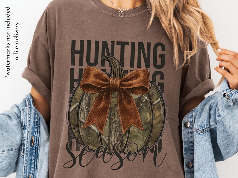 May include: Brown t-shirt with a pumpkin design. The pumpkin has a camouflage pattern and a large brown bow. The words "HUNTING SEASON" are printed above and below the pumpkin in black and cursive font.