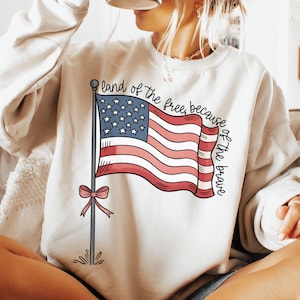 以下が含まれることがあります： 赤、白、青のアメリカ国旗のグラフィックと「land of the free, because of the brave」というテキストが書かれた白いスウェットシャツ。