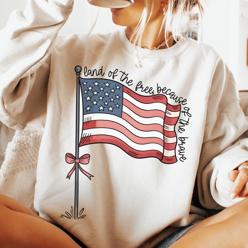 以下が含まれることがあります： 赤、白、青のアメリカ国旗のグラフィックと「land of the free, because of the brave」というテキストが書かれた白いスウェットシャツ。