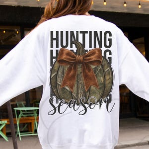 May include: White sweatshirt with a pumpkin graphic. The pumpkin has a camouflage pattern and a brown bow. The words "HUNTING SEASON" are printed above and below the pumpkin in a black font.