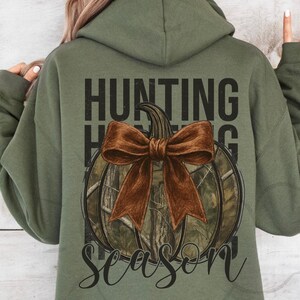 May include: Olive green hoodie with a graphic on the back. The graphic features a pumpkin with a brown bow and camouflage pattern, with the words "HUNTING SEASON" in black.