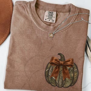 May include: A brown Comfort Colors t-shirt featuring a pumpkin design with a camouflage pattern and a brown bow. A silver necklace with a small pendant rests on the shirt. A brown hat is visible in the upper left corner.