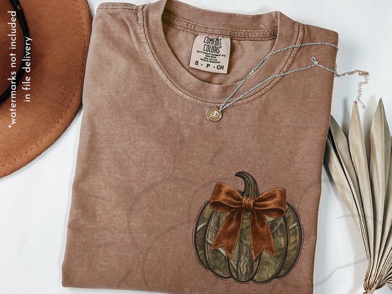 May include: A brown Comfort Colors t-shirt featuring a pumpkin design with a camouflage pattern and a brown bow. A silver necklace with a small pendant rests on the shirt. A brown hat is visible in the upper left corner.