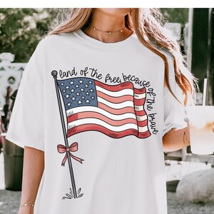 以下が含まれることがあります： 赤、白、青のアメリカ国旗のグラフィックと「land of the free, because of the brave」というテキストが書かれた白いTシャツ。
