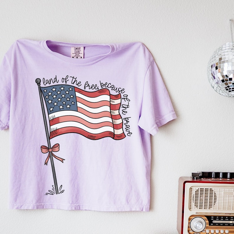 以下が含まれることがあります： アメリカの国旗と「land of the free, because of the brave」というテキストが描かれたパープルのTシャツです。