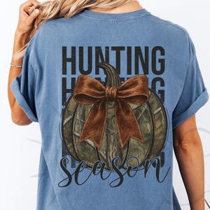 May include: A blue t-shirt with a pumpkin design. The pumpkin has a camouflage pattern and a brown bow. The words "HUNTING SEASON" are printed above and below the pumpkin in black.