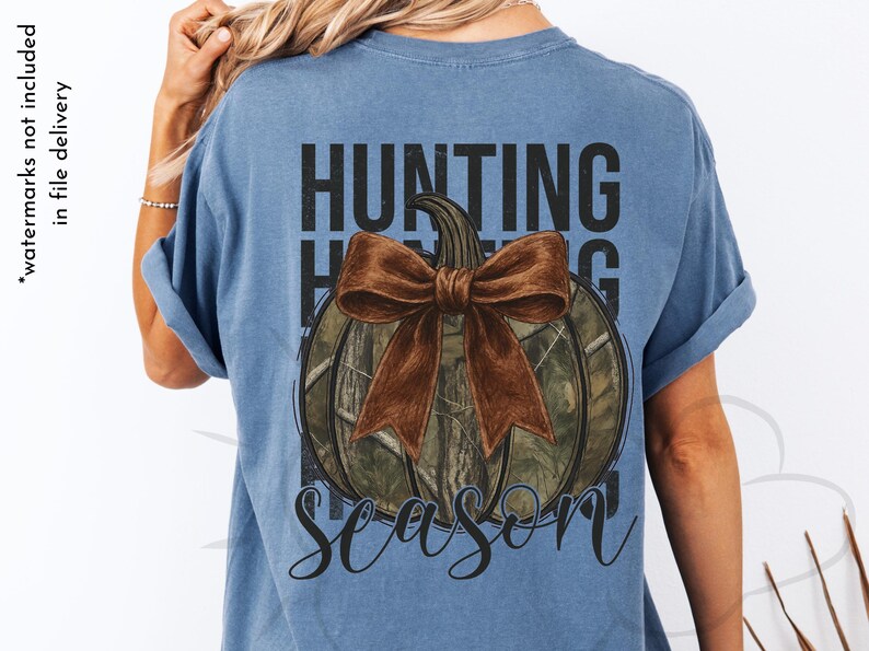 May include: A blue t-shirt with a pumpkin design. The pumpkin has a camouflage pattern and a brown bow. The words "HUNTING SEASON" are printed above and below the pumpkin in black.