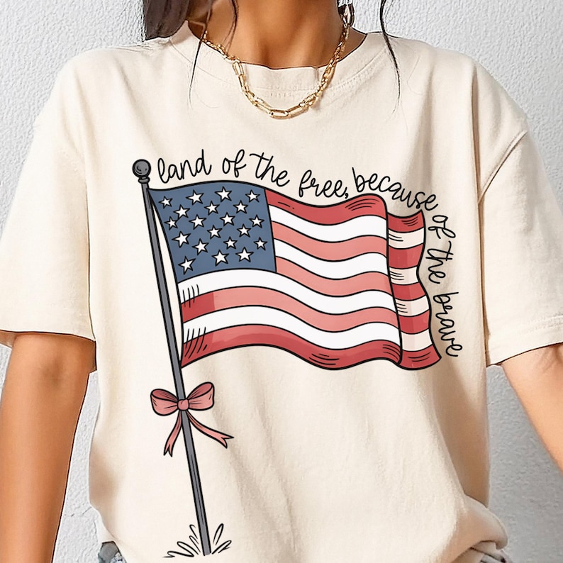 以下が含まれることがあります： アメリカの国旗と「land of the free, because of the brave」というテキストが描かれたベージュのTシャツです。