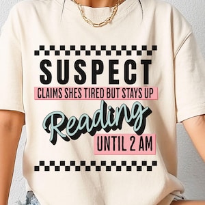 Op de afbeelding: Een wit T-shirt met een zwart-wit geruit patroon aan de boven- en onderkant. De tekst "SUSPECT" is in zwart, en de tekst "CLAIMS SHES TIRED BUT STAYS UP Reading UNTIL 2 AM" is in roze en blauw.