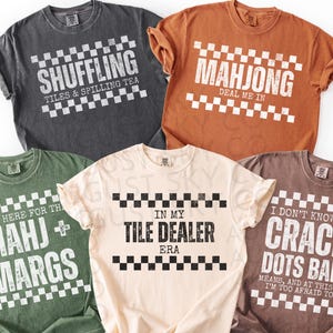 Puede incluir: Cinco camisetas de varios colores, cada una con un diseño único relacionado con el Mahjong. Las camisetas presentan frases como "Shuffling Tiles & Spilling Tea" y "Mahjong Deal Me In" en un estilo de tablero de ajedrez.