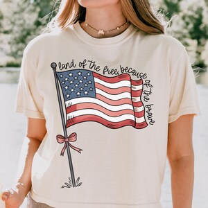 以下が含まれることがあります： クリーム色のTシャツに、赤、白、青のアメリカ国旗のグラフィックと「land of the free, because of the brave」のテキストがプリントされています。