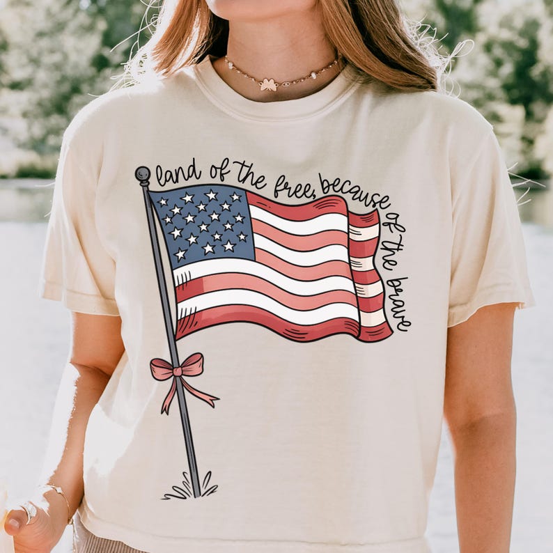 以下が含まれることがあります： クリーム色のTシャツに、赤、白、青のアメリカ国旗のグラフィックと「land of the free, because of the brave」のテキストがプリントされています。