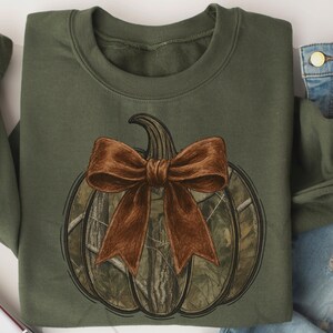 May include: Olive green sweatshirt featuring a pumpkin design with a brown bow. The pumpkin has a camouflage pattern. The design is centered on the front of the sweatshirt. The sweatshirt is folded, with a pair of blue jeans visible.