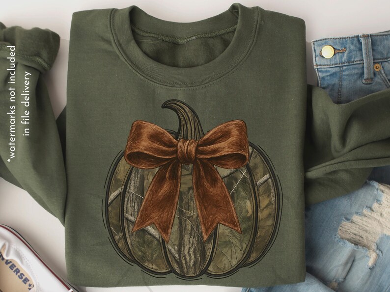 May include: Olive green sweatshirt featuring a pumpkin design with a brown bow. The pumpkin has a camouflage pattern. The design is centered on the front of the sweatshirt. The sweatshirt is folded, with a pair of blue jeans visible.