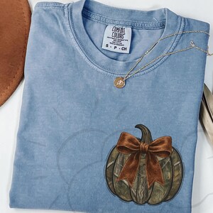 May include: A light blue Comfort Colors t-shirt with a pumpkin graphic. The pumpkin has a camouflage pattern and a brown bow. A gold necklace with a pendant is draped across the shirt.