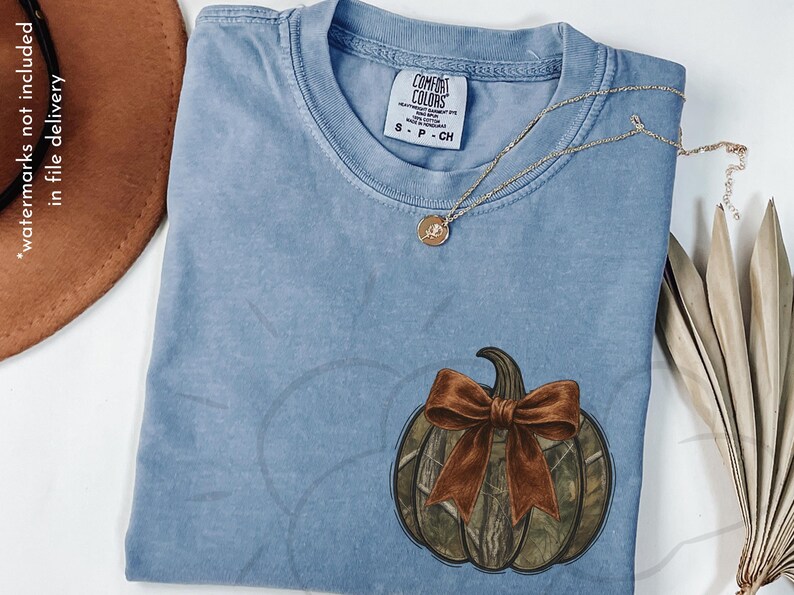 May include: A light blue Comfort Colors t-shirt with a pumpkin graphic. The pumpkin has a camouflage pattern and a brown bow. A gold necklace with a pendant is draped across the shirt.