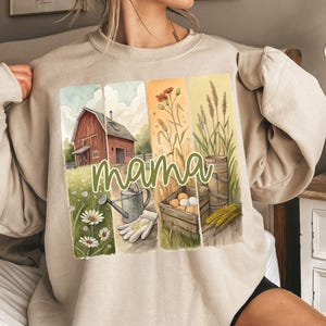 Może przedstawiać: Beżowa bluza z akwarelową ilustracją wiejskiego domu, kwiat&oacute;w, pszenicy i konewki. Tekst "mama" jest napisany zielonym, ozdobnym pismem na przodzie bluzy.