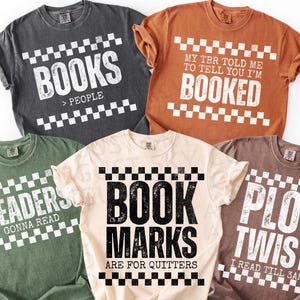 May include: Five short-sleeved t-shirts in various colours, each with a book-related quote in white block letters. The shirts are grey, orange, green, cream, and brown. The text includes "BOOKS > PEOPLE", "MY TBR TOLD ME TO TELL YOU I'M BOOKED", "READERS GONNA READ", "BOOK MARKS ARE FOR QUITTERS", and "PLOT TWIST I READ TILL 3AM".