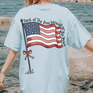以下が含まれることがあります： 薄い青色のTシャツで、背面に赤、白、青のアメリカ国旗のグラフィックが描かれています。旗は風になびいており、ポールにピンクのリボンが結ばれています。旗の周りに「land of the free, because of the brave」というテキストが書かれています。