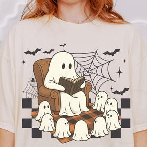 May include: Cream-colored t-shirt featuring a cartoon ghost reading a book in a brown armchair, surrounded by smaller ghosts. The design includes bats, a spiderweb, and a checkered rug, creating a Halloween theme.