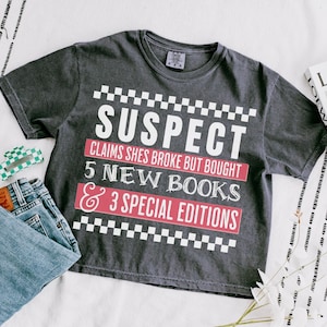 Może przedstawiać: Szary t-shirt z białą kratką i napisem "SUSPECT CLAIMS SHES BROKE BUT BOUGHT 5 NEW BOOKS & 3 SPECIAL EDITIONS" w kolorze białym na czerwonym tle.