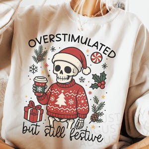 May include: Cream-colored sweatshirt with a festive skeleton graphic. The skeleton wears a Santa hat and a red sweater with a Christmas tree design. Text reads "OVERSTIMULATED but still festive". The design includes snowflakes, holly, and a coffee cup.