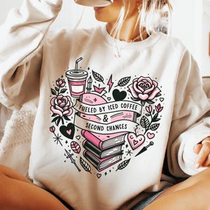 Pode incluir: Camisola branca com um design de coração rosa com uma chávena de café, livros e o texto "Fueled by Iced Coffee & Second Chances".