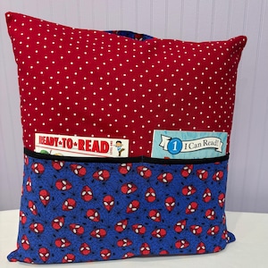 May include: A reading pillow with a red polka dot top and a blue bottom patterned with red Spider-Man faces and webs. The pillow has a black pocket holding two children's books. The books read "Ready to Read" and "I Can Read!"