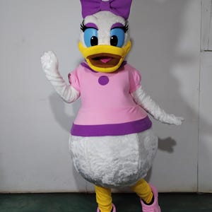 Könnte beinhalten: Ein weißes Enten-Maskottchen-Kostüm mit einem rosa Kleid, einer lila Schleife und gelben Schuhen. Das Kostüm hat einen gelben Schnabel und blaue Augen.