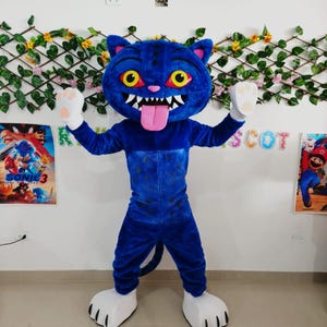 以下が含まれることがあります： 大きな頭、黄色い目、ピンクの舌を持つ青い猫のマスコットコスチューム。コスチュームは白い手と足を持っています。背景にはポスターと装飾的な緑があります。