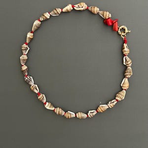 Puede incluir: Un collar con pequeñas conchas marinas estampadas ensartadas en un cordón rojo. El collar tiene un cierre dorado y una sección de coral rojo. Las conchas son de color beige y marrón, creando una estética natural y playera.