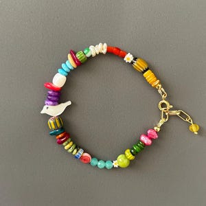Può includere: Un braccialetto di perline colorate con una varietà di forme e dimensioni. Il braccialetto presenta un ciondolo a forma di uccello bianco e una chiusura color oro. Le perline includono rosso, giallo, verde, blu e rosa. Il braccialetto è lungo circa 18 cm.