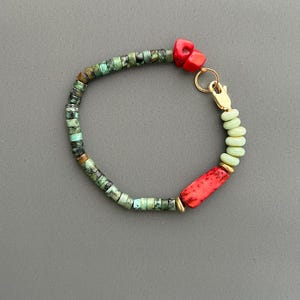 Peut inclure: Un bracelet en perles turquoise, vert et rouge avec un fermoir doré et une petite perle dorée. Le bracelet est fait de pierres naturelles et a un style bohème.