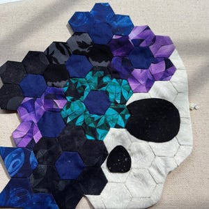 May include: A skull-shaped fabric applique constructed from numerous small hexagonal pieces of cloth. The design features a white fabric base for the skull, with black fabric eye and nose sockets, and a mosaic of blue, purple, and teal hexagons.