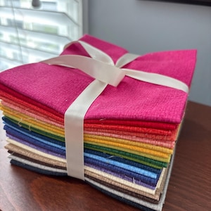May include: A stack of colorful fabric squares, neatly arranged and tied with a cream-colored ribbon. The fabrics display a spectrum of colors, from vibrant pink to deep navy and brown, suggesting a quilting or crafting project.