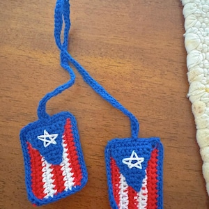 Peut inclure: Deux ornements rectangulaires au crochet arborant le motif du drapeau de Porto Rico avec des rayures rouges et blanches, un triangle bleu et une étoile blanche brodée sur chacun. Les deux pièces sont reliées par une boucle en crochet bleu.