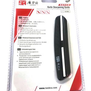 K&ouml;nnte beinhalten: Schwarzer Messersch&auml;rff&uuml;hrer in einer transparenten Kunststoffverpackung. Der F&uuml;hrer hat einen wei&szlig;en Streifen und den Text "Knife Sharpening Guide". Die Verpackung enth&auml;lt Text in mehreren Sprachen, darunter Englisch, Chinesisch und Deutsch.
