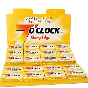 May include: A yellow cardboard display box filled with Gillette 7 O'Clock SharpEdge razor blade packs. The display features the Gillette logo and product name in red and black. Each pack is yellow with red and black text.