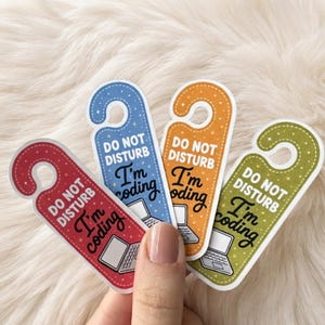 Op de afbeelding: Vier deurhangers in rood, blauw, oranje en groen, elk met witte tekst DO NOT DISTURB en zwarte tekst I'm coding, vergezeld van een kleine witte laptopillustratie en een patroon van kleine witte sterretjes.