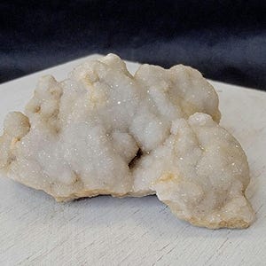 May include: A cluster of druzy quartz crystals, showcasing a sparkling, crystalline surface. The quartz has a creamy white color with hints of yellow and sits on a white surface. The crystals have a rough, uneven texture.