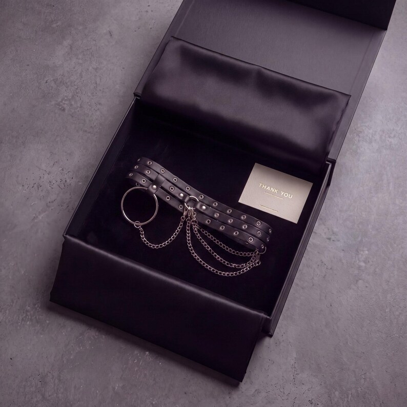 May include: A black choker with silver-toned eyelets, a large circular ring, and three draped chains, presented in a black gift box with a small card that reads THANK YOU. The choker features multiple rows of metal hardware and a secure clasp.