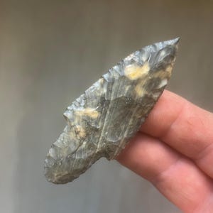 May include: A hand-held, gray and tan arrowhead with a sharp point and notched base. The stone tool has a layered appearance with variations in color and texture, suggesting it is made from flint or a similar material.