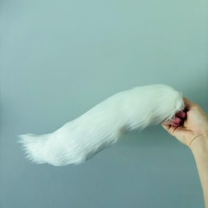 May include: A long, plush, white faux fur tail accessory. The tail features thick, soft, synthetic white fibers that taper toward the end, creating a realistic animal-like appearance with a dense, fluffy texture throughout the entire length.