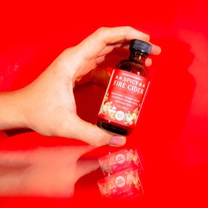 May include: A small glass bottle of "Spicy Fire Cider" with a black cap and a red label. The label includes text and illustrations of fruits and vegetables. Two smaller labels are reflected below the bottle.