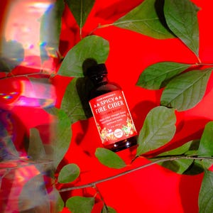 May include: A bottle of "Spicy Fire Cider" with a red and white label, surrounded by green leaves, rests on a vibrant red background. The label features text and illustrations. The bottle has a black cap. The image is well-lit and the colors are vivid.