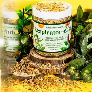 Puede incluir: Un frasco de vidrio de té de hierbas "Respirator-ease" para el apoyo respiratorio, con tapa blanca y etiqueta con ilustraciones botánicas. El frasco está rodeado de vegetación y descansa sobre una rodaja de madera. El frasco contiene 46 gramos de té.