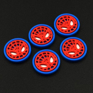 以下が含まれることがあります： 白いクモの巣のデザインが施された、赤と青の丸いボタンが5つ。クモの巣のデザインの中央には白いクモの顔があります。