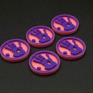 May include: Five round, two-tone tokens with a handprint design. Each token has a pink center with a raised purple handprint and a purple border. The tokens are arranged on a dark surface.