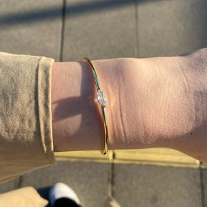May include: A gold-colored bangle bracelet with a marquise-cut clear gemstone. The bracelet is thin and simple, with the gemstone set in the center. The jewelry is worn on a wrist.